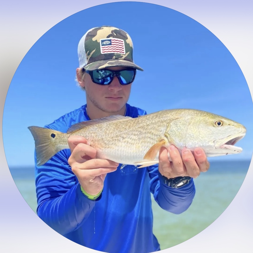 Capt. Gavin Ward in a camo cap and sunglasses holding a redfish in the shallows