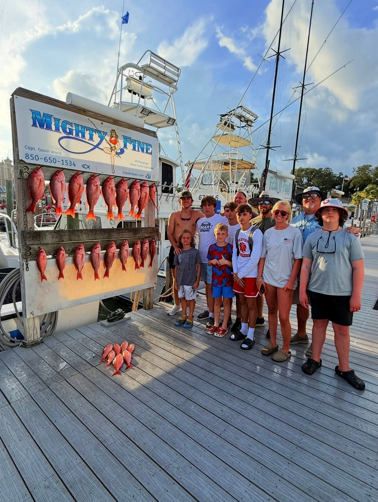 A big multi-generation family group at the dock in golden-hour light beside the Mighty Fine catch board full of red snapper